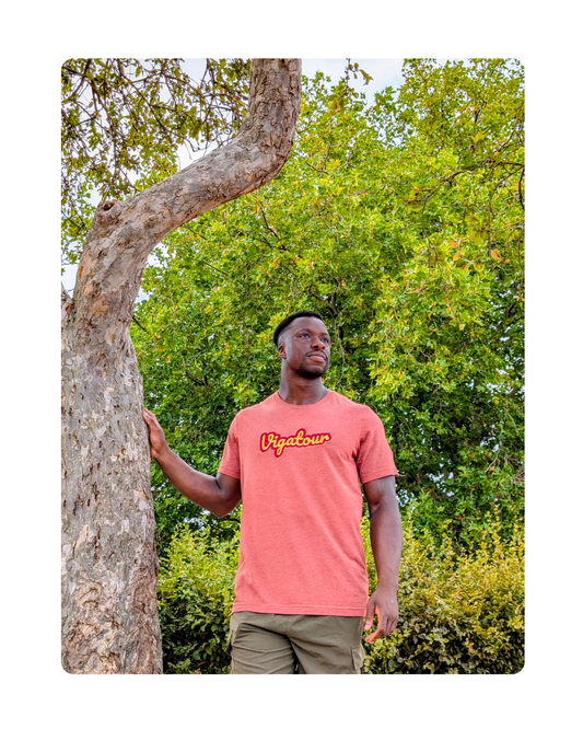 A man wearing a red t-shirt with the yellow and red 'Vigatour' writing embroidered on the chest, standing outdoors next to a tree with green foliage in the background.