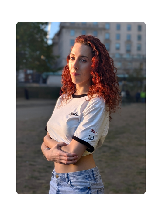 A woman with curly red hair wearing a white ringer top with black trim and light blue jeans, standing outdoors in a park-like setting with a building in the background. They are wearing the Vigatour 'Der Klassiker' shirt.